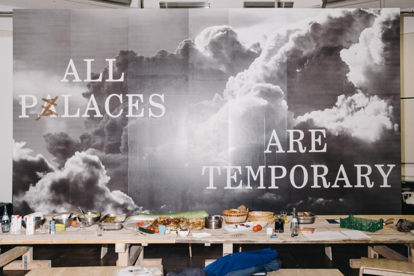 Ein großer Schriftzug „All Palaces Are Temporary“ prangt über einem mit Essen bedeckten Holztisch in einem Ausstellungsraum.