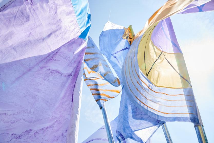 Das Bild zeigt eine Gruppe von Menschen im Freien vor farbigen Fahnen, die in einer Wiese zwischen modernen Gebäuden aufgestellt sind.