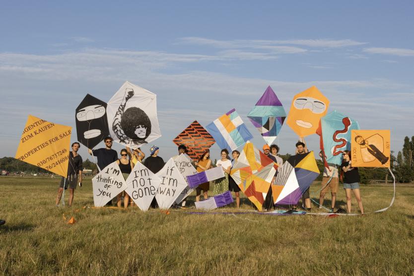 Das Bild zeigt eine Gruppe von Menschen auf einer Wiese, die selbstgestaltete Drachen mit politischen und grafischen Motiven in die Kamera halten.