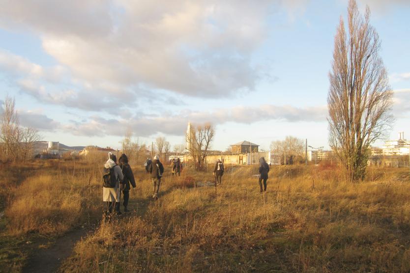 Mehrere Personen gehen bei tiefstehender Sonne über ein brachliegendes, wild bewachsenes Gelände. Die Szene wirkt ruhig und gemeinschaftlich, mit weitem Blick auf die winterliche Landschaft und vereinzelte Gebäude im Hintergrund. Die Menschen sind warm gekleidet und bewegen sich in kleinen Gruppen.