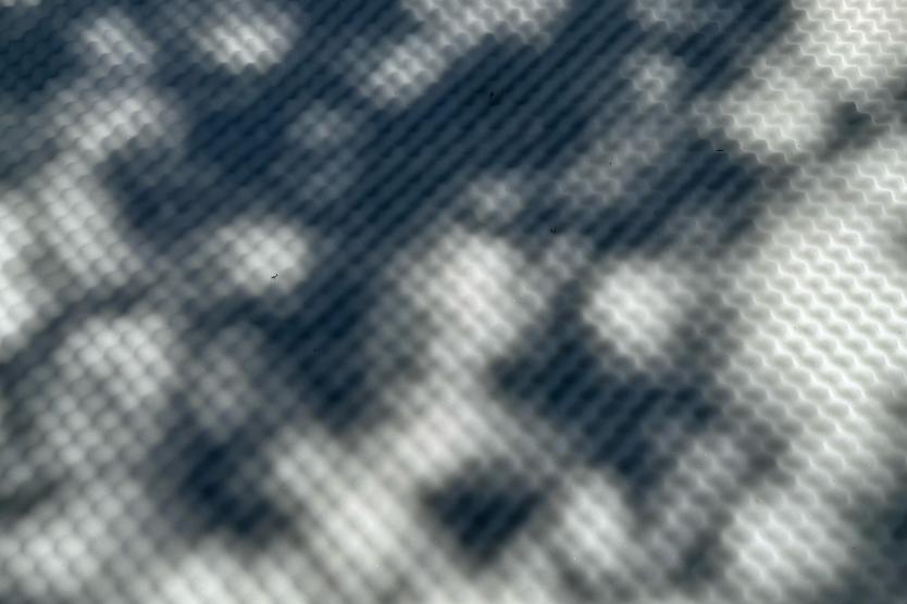 Licht und Schatten werfen ein rasterartiges Muster auf eine strukturierte Oberfläche und erzeugen ein abstraktes Spiel aus Formen und Tiefen.