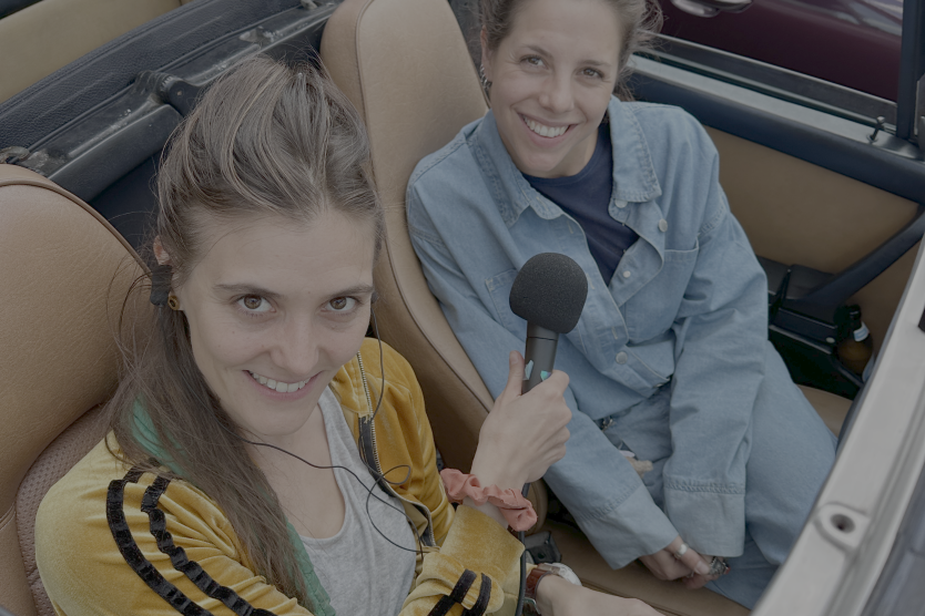 Zwei Frauen sitzen in einem offenen Cabrio, die eine hält ein Mikrofon, beide schauen fröhlich in die Kamera.