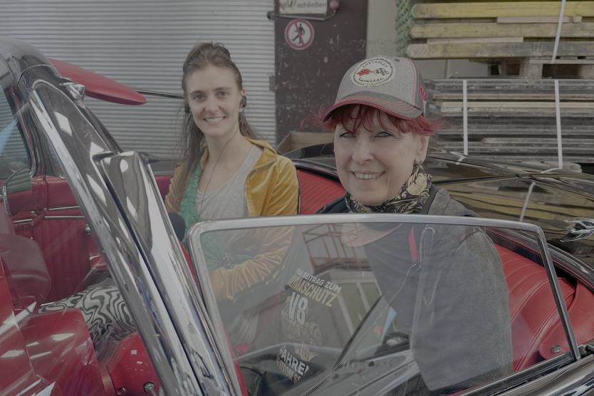 Zwei Frauen sitzen lächelnd in einem klassischen Cabrio mit roter Innenausstattung in einer Werkhalle.