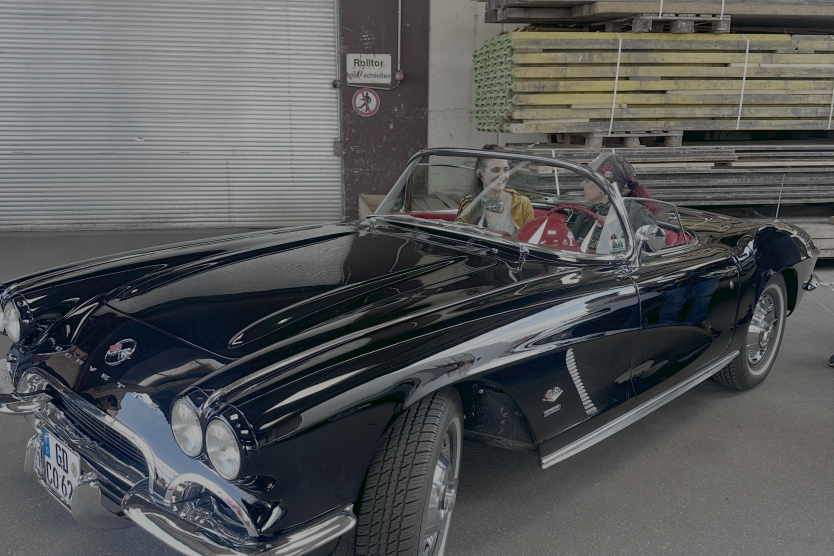 Zwei Frauen sitzen in einem schwarzen Oldtimer-Cabrio mit rotem Innenraum in einer Halle vor gestapelten Paletten.