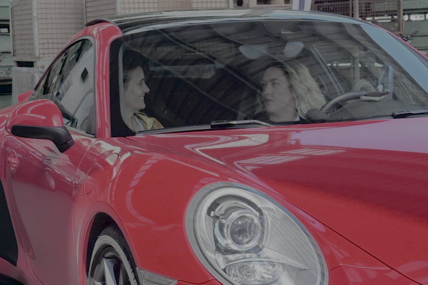 Zwei Frauen führen ein Gespräch in einem roten Sportwagen vor einer Industrie-Kulisse mit Metallkisten und Holzpaletten.