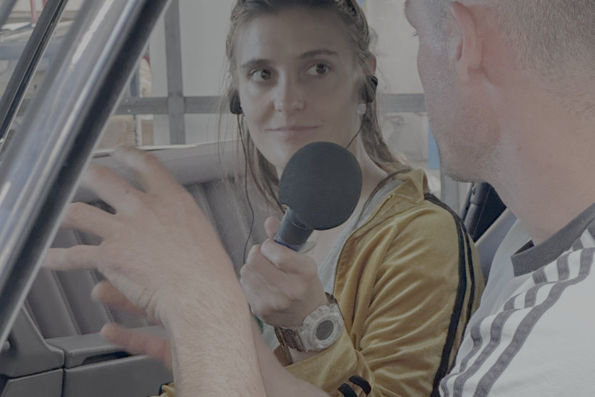 Eine Frau mit Mikrofon interviewt einen Mann in einem Auto, beide wirken konzentriert im Gespräch.