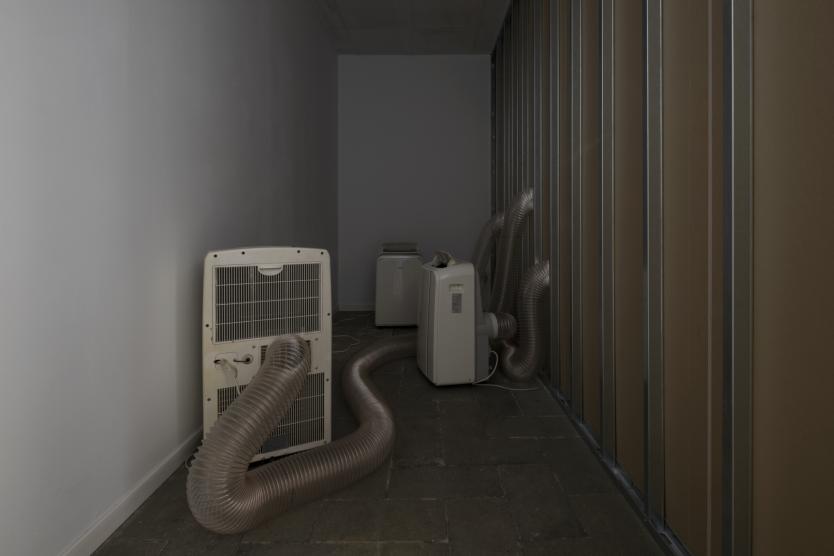 In einem schmalen, düsteren Raum stehen mehrere mobile Klimageräte, deren dicke Schläuche sich wie Tentakel über den Boden und an der Wand entlangziehen.“  Wenn du mir deinen ursprünglichen Satz nennst, kann ich die Alternative noch gezielter auf deinen Stil abstimmen.