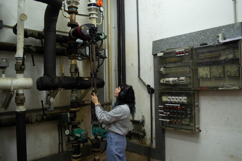 Eine Frau mit Kopfhörern steht in einem Technikraum vor alten Rohrleitungen und Schaltkästen, während sie mit einem Mikrofon auf einem Stativ aufmerksam die Geräusche der Maschinen aufnimmt.