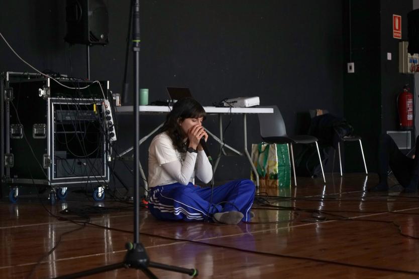 Eine junge Frau sitzt mit geschlossenen Augen auf dem Boden eines Studios und hält ein Mikrofon mit beiden Händen, während sie sich auf den Klang konzentriert – umgeben von Kabeln, Technik und minimalistischer Ausstattung.
