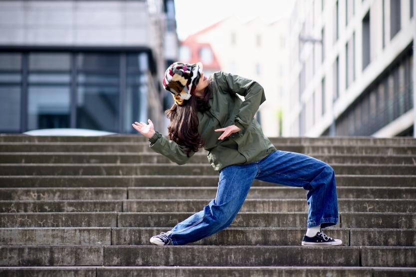 Person tanzt in dynamischer Pose auf einer Außentreppe vor modernen Gebäuden.