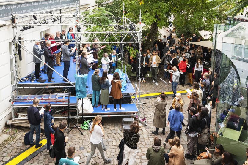 Ein Chor singt auf einer provisorischen Bühne im Freien, während zahlreiche Menschen im Hof zuhören und mitmachen.