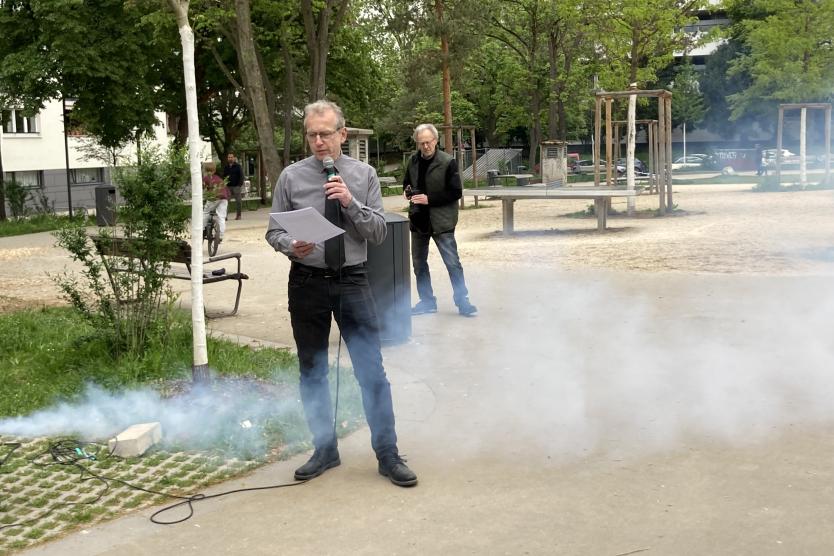 Ein Redner hält im Park eine Ansprache, während im Hintergrund Nebel aufzieht und eine zweite Person neben dem Lautsprecherpult steht.