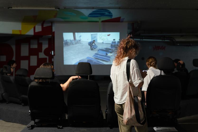 Menschen sitzen in Autositzen und schauen einen Film auf einer Leinwand. Die Atmosphäre ist ruhig, der Raum wirkt wie eine ungewöhnliche Kinoumgebung.