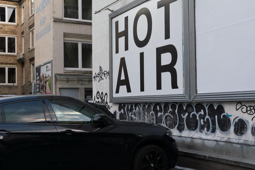Ein schwarzes Auto steht vor einer Hauswand mit Graffiti. An der Wand hängt ein großes Schild mit der Aufschrift „HOT AIR“.