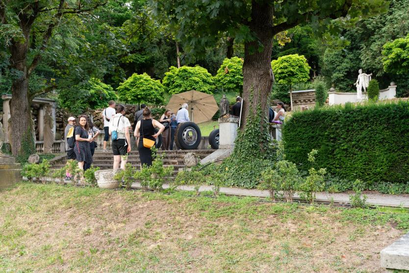 Menschen versammeln sich im Grünen bei einer Treppe mit alten Skulpturen. Zwischen Bäumen und Hecken stehen große Reifen und ein Sonnenschirm.
