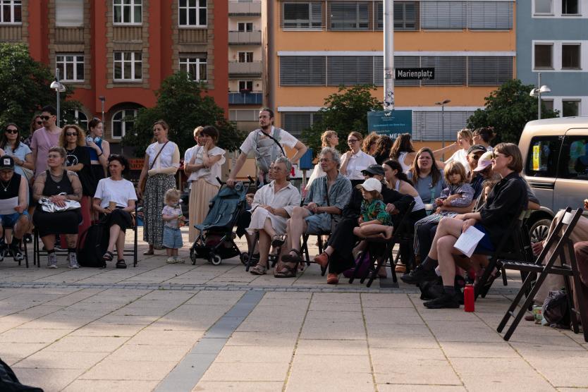 Eine Gruppe von Menschen sitzt und steht auf einem öffentlichen Platz. Einige hören zu, andere unterhalten sich. Kinder sind ebenfalls dabei. Im Hintergrund ist ein Schild mit der Aufschrift „Marienplatz“ zu sehen.