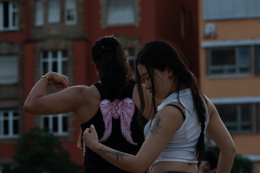 Zwei Menschen tanzen draußen vor einem Gebäude. Eine Person trägt ein schwarzes Top mit rosa Engelsflügeln auf dem Rücken.