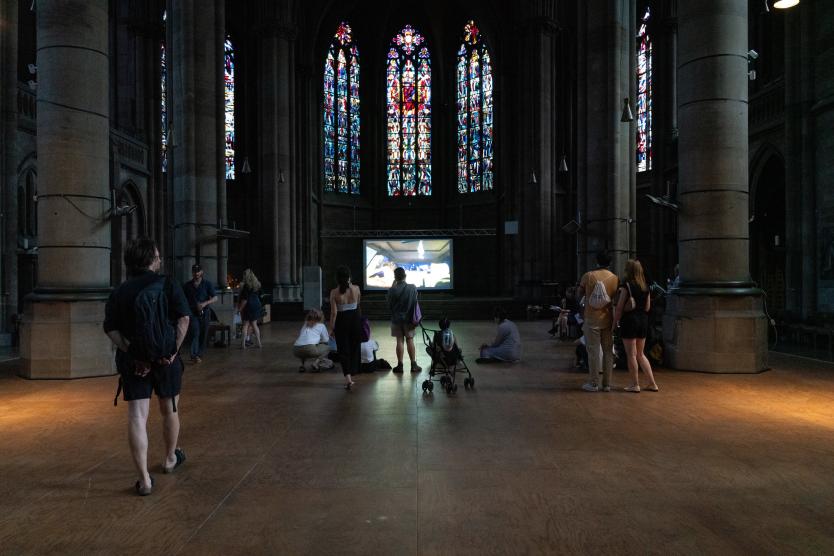 Menschen stehen und sitzen in einer Kirche mit bunten Fenstern. Auf einer Leinwand läuft ein Film. Die Stimmung ist ruhig und konzentriert.