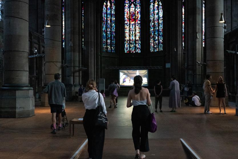 Menschen gehen durch das Kirchenschiff in Richtung einer Leinwand. Bunte Fenster werfen Licht in den dunklen Raum. Die Szene wirkt ruhig und aufmerksam.