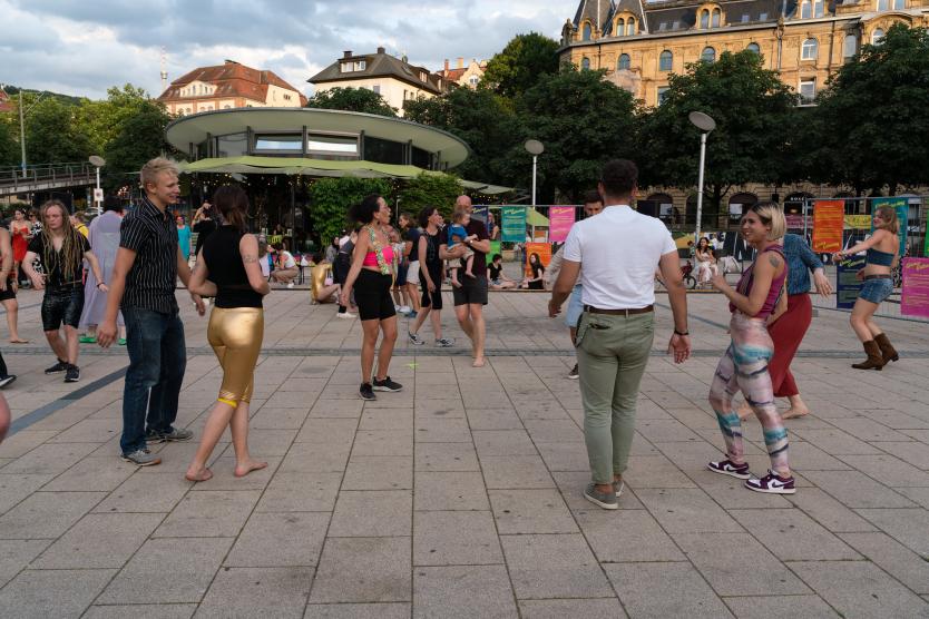 Menschen tanzen gemeinsam auf einem offenen Platz. Die Stimmung wirkt lebendig und offen, im Hintergrund sind bunte Plakate zu sehen.