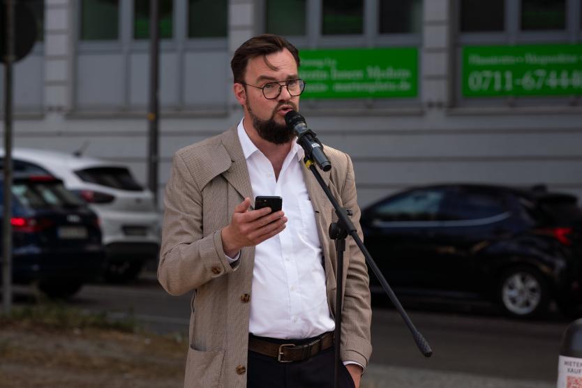 Eine Person spricht draußen in ein Mikrofon und liest dabei von einem Handy. Im Hintergrund sind parkende Autos und ein Gebäude mit grünen Schildern zu sehen. Es handelt sich um das Grußwort Marc Gegenfurtner, Amtsleiter Kulturamt Stadt Stuttgart.