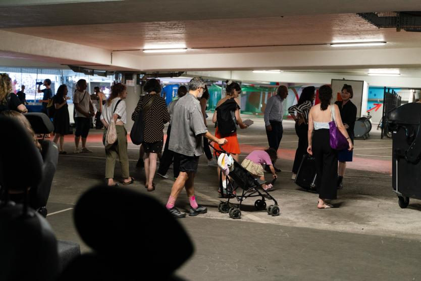 Menschen bewegen sich durch ein Parkhaus. Einige unterhalten sich, andere schieben Kinderwagen oder schauen sich um.