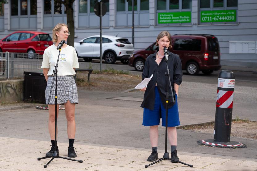 Zwei Personen stehen mit Mikrofonen auf einem Platz. Eine liest von einem Zettel, die andere schaut zu. Autos und Häuser sind im Hintergrund zu sehen.