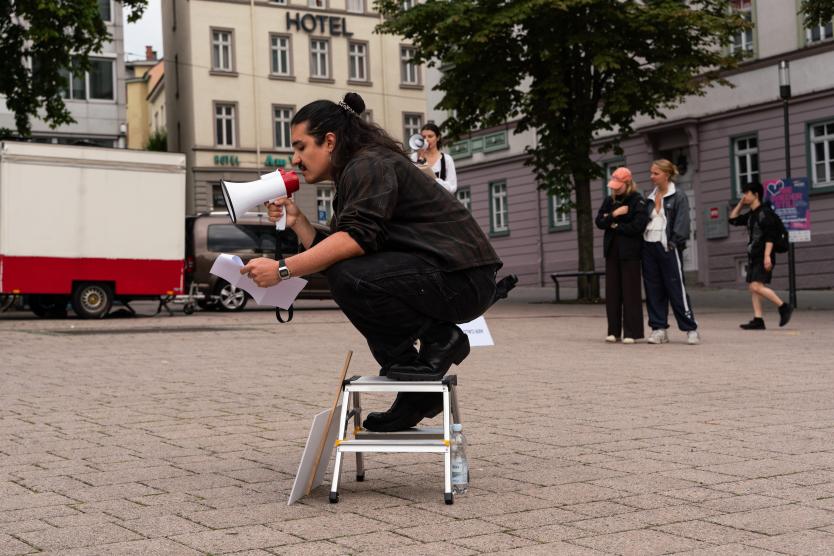 Eine Person hockt auf einem kleinen Klapptritt auf einem städtischen Platz und spricht mit einem Megafon in der Hand in Richtung einer kleinen Gruppe Zuhörender, während ein weiteres Megafon und Informationsmaterial sichtbar sind.