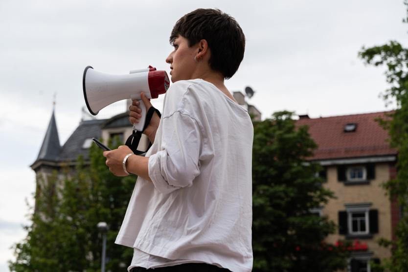 Eine Person steht im Freien mit einem Megafon in der einen Hand und einem Smartphone in der anderen, während im Hintergrund Gebäude und Bäume zu sehen sind.