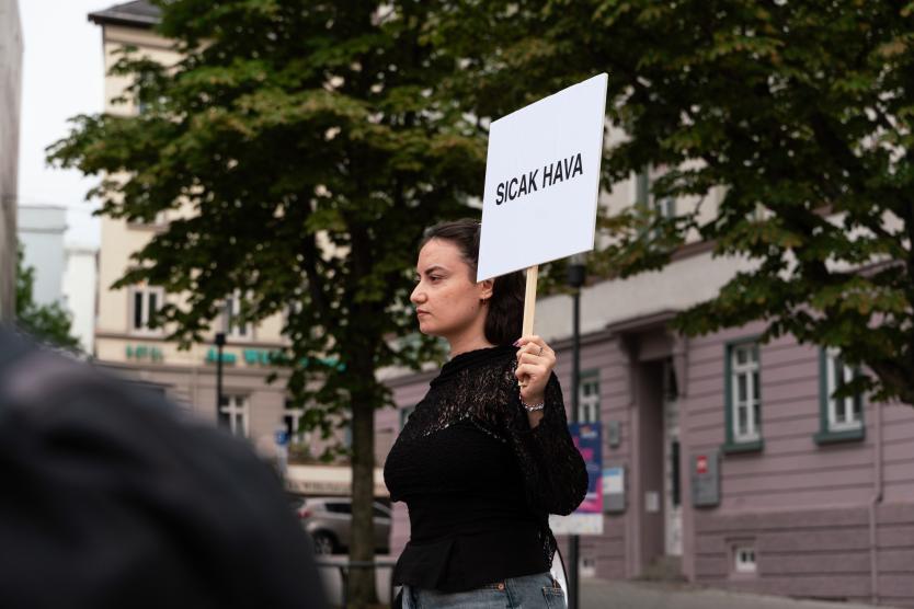 Eine Person steht im Freien mit einem Megafon in der einen Hand und einem Smartphone in der anderen, während im Hintergrund Gebäude und Bäume zu sehen sind.