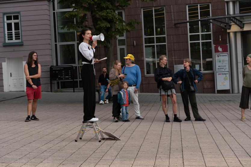 Eine Person steht auf einem Klapptritt und spricht mit einem Megafon zu einer Gruppe umstehender Menschen auf einem städtischen Platz, während ein Schild auf dem Boden liegt.