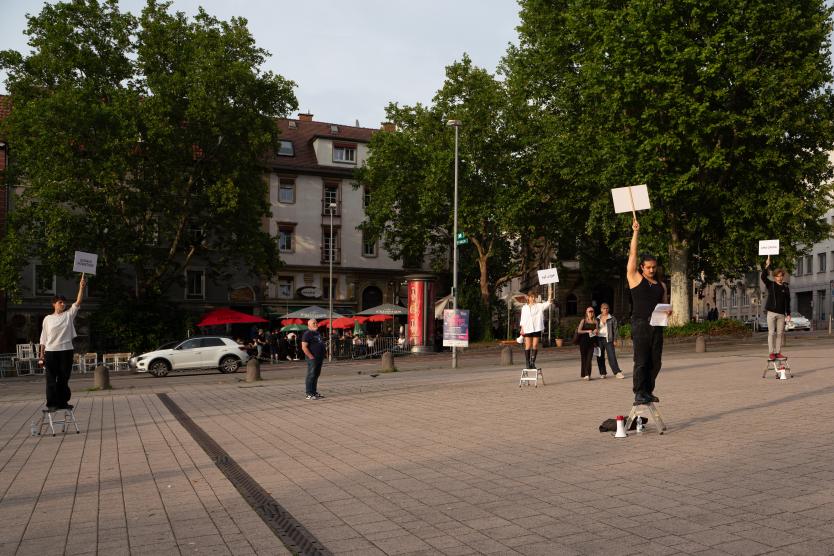 Mehrere Personen stehen mit erhobenen Schildern in verschiedenen Sprachen auf kleinen Tritten verteilt über einen öffentlichen Platz, während Megafone und Materialien auf dem Boden liegen und Passant*innen im Hintergrund vorbeigehen.