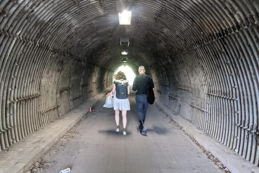 Zwei Personen durchqueren einen schmalen, geriffelten Fußgängertunnel – ein alltäglicher Raum, der durch O.J.A.I.s Intervention zum Ort spekulativer Infrastrukturpoesie wird.