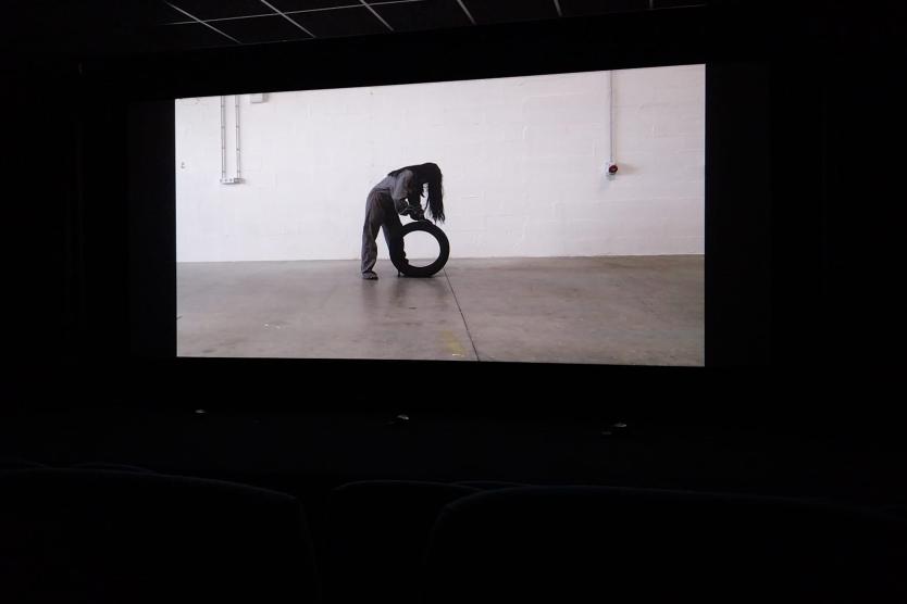 Eine Videoaufnahme in einem Kinosaal zeigt eine Person in grauer Kleidung, die sich in einem leeren Raum mit weißer Wand über einen Autoreifen beugt.