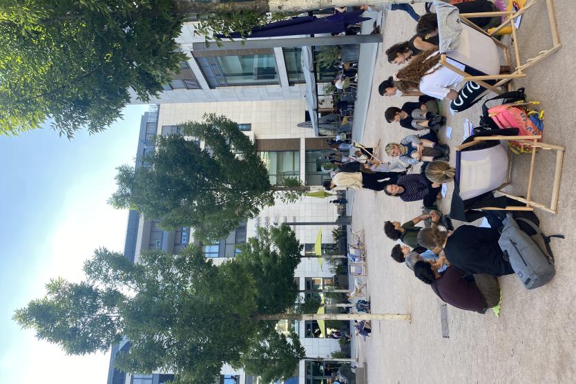 Eine Gruppe von Menschen sitzt im Kreis auf einem öffentlichen Platz, umgeben von Bäumen und moderner Architektur. Vor ihnen liegen Papiere und Gegenstände auf dem Boden. Einige Teilnehmende sitzen auf bunten Matten oder in Liegestühlen.
