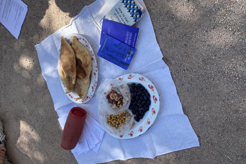 Auf einer weißen Decke, die auf dem Boden ausgebreitet ist, liegen zwei Teller mit Gebäck, Oliven, Nüssen und getrockneten Früchten. Daneben befinden sich mehrere Bücher und ein roter Lautsprecher. Um die Decke herum sind lose Blätter und Papiere verteilt.