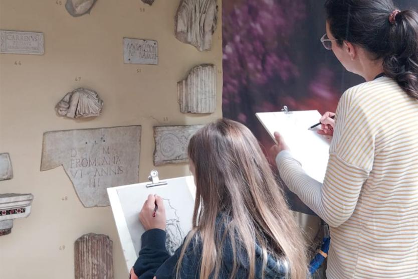 Zwei Personen zeichnen konzentriert antike Relieffragmente an einer Museumswand.