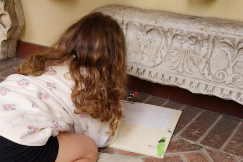 Ein Kind liegt auf dem Boden und zeichnet konzentriert ein antikes Relief.