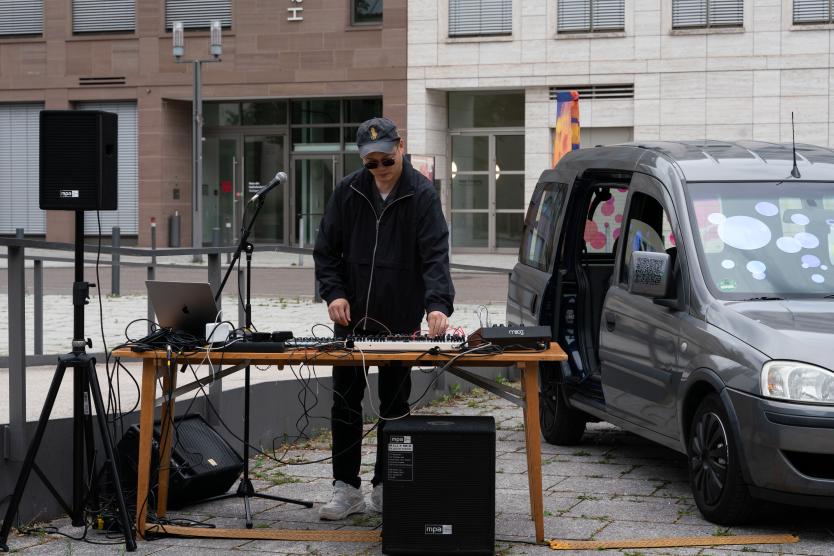 Ein Performer mit Cap und Sonnenbrille bedient ein Mischpult im Freien. Der Tisch ist voll verkabelt, Lautsprecher und ein Mikrofon stehen bereit. Daneben ein geparktes Auto mit bunten Kreisen auf den Fenstern – eine mobile Bühne im Stadtraum.