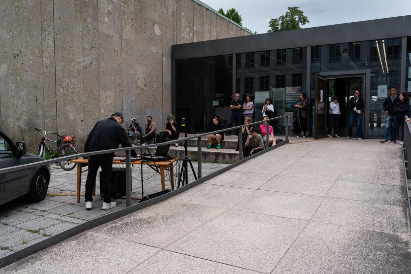 Ein Musiker steht an einem Tisch mit elektronischem Equipment, der direkt vor einem modernen Gebäude aufgebaut ist. Um ihn herum hat sich ein kleines Publikum versammelt – einige sitzen auf den Stufen, andere stehen an der Rampe oder im Eingangsbereich. Im Vordergrund ist der Rand eines Autos zu sehen, im Hintergrund Fahrräder und Stadtarchitektur.