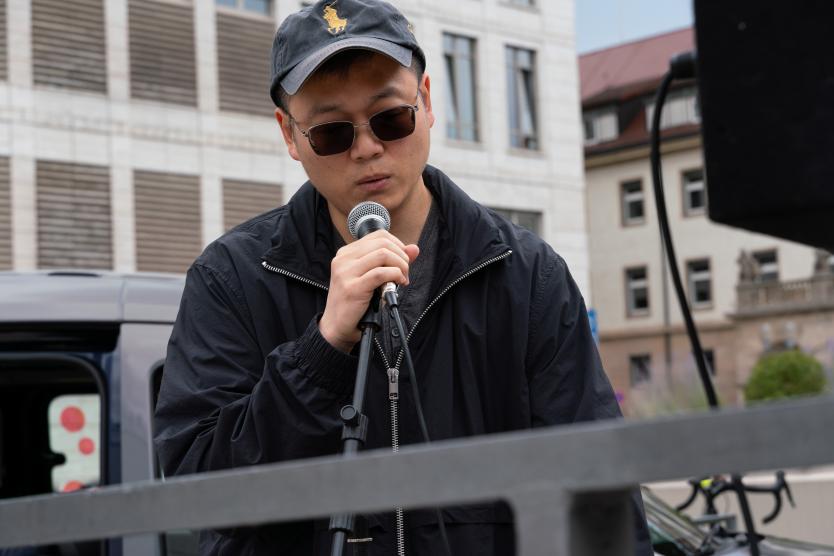 Ein Musiker steht in Nahaufnahme am Mikrofon. Er trägt eine dunkle Jacke, Sonnenbrille und eine Kappe. Die Szene spielt sich draußen ab, vor urbanem Hintergrund mit modernen und historischen Gebäuden.