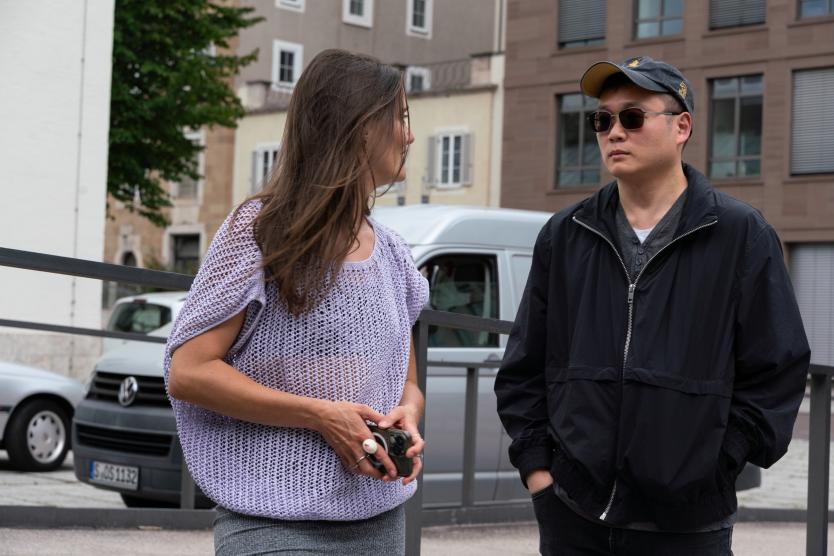 Zwei Personen stehen im urbanen Außenraum im Gespräch nebeneinander. Die Frau trägt ein gestricktes, fliederfarbenes Oberteil und hält ein Smartphone in der Hand. Der Mann trägt eine dunkle Jacke, Basecap und Sonnenbrille. Im Hintergrund sind Autos, Gebäude und ein Gehweg zu sehen.
