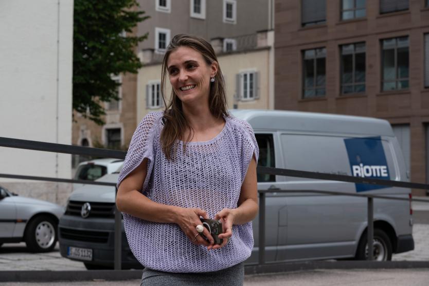 Eine Frau im gestrickten Oberteil lächelt, hält ein Smartphone in der Hand. Im Hintergrund: Stadt, Autos, Transporter mit „RIOTTE“-Schriftzug.