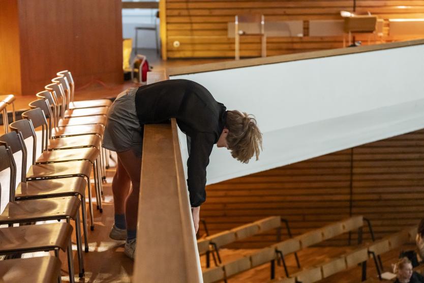 Eine Performerin lehnt mit dem Oberkörper über die Brüstung einer Empore, die Arme schlaff nach unten hängend. Ihr Blick ist gesenkt, der Körper wirkt entspannt und schwer. Der Hintergrund zeigt leere Stuhlreihen und die Holzverkleidung des Raums, der in warmes Licht getaucht ist.
