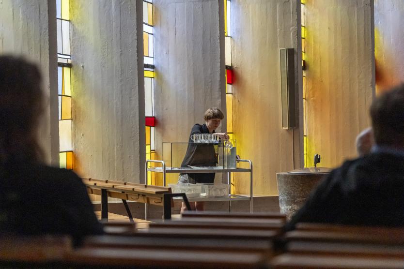 Eine Person bereitet an einem Wagen mit Gläsern und Wasser in der Kirche etwas vor. Farbige Fenster tauchen die Szene in warmes Licht.