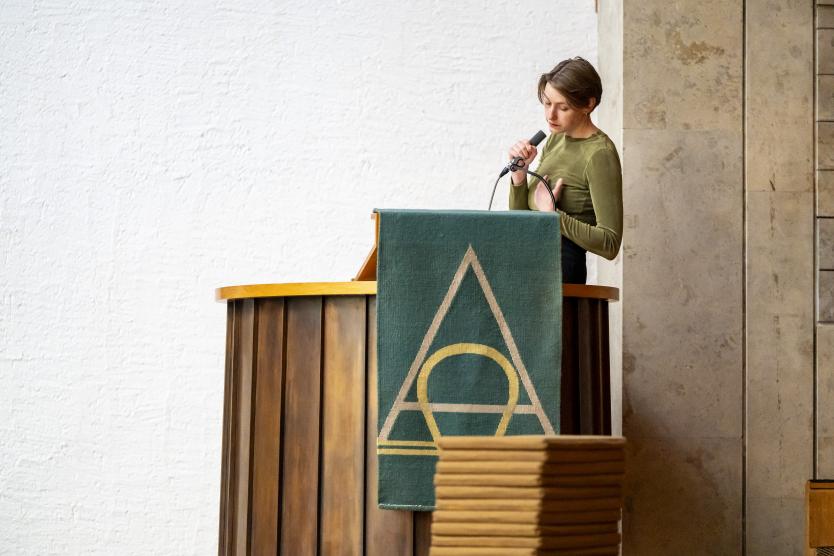Eine Person steht am Lesepult in einer Kirche und spricht in ein Mikrofon. Vor ihr hängt ein grünes Tuch mit christlichen Symbolen.