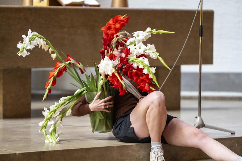 Eine Person sitzt auf einer Kirchenstufe und umarmt eine große Vase mit weißen und roten Gladiolen, die ihr Gesicht fast vollständig verdecken.