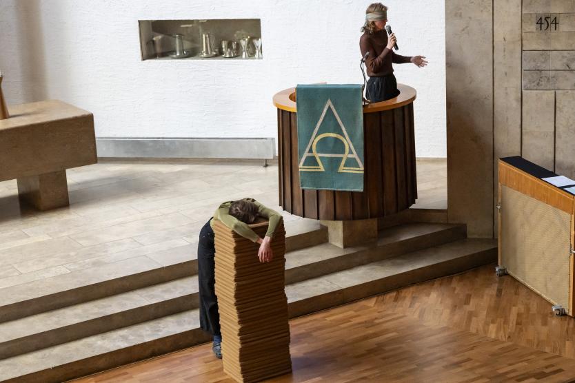 Eine Performerin mit Augenbinde spricht von der Kanzel, während eine andere erschöpft über einem Stapel Kniekissen hängt.