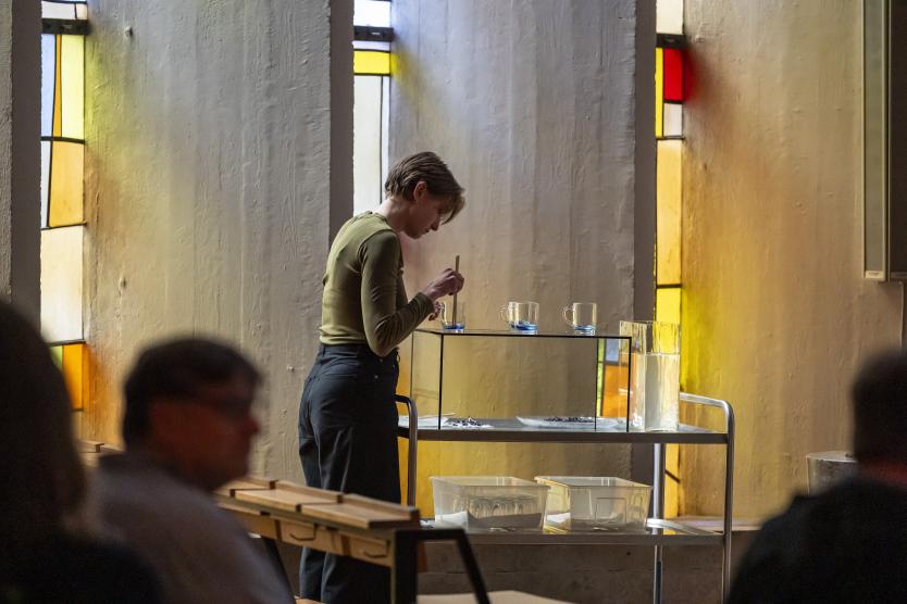 Eine Performerin bereitet konzentriert Gläser mit blauer Flüssigkeit vor – ein stiller Moment ritueller Handlung im farbig durchleuchteten Kirchenraum.