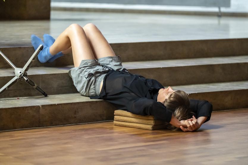 Eine Performerin liegt entspannt auf dem Boden, den Kopf auf Kissen gebettet – ein Moment der Stille und Kontemplation im sakralen Raum.